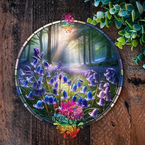 May include: A stained glass round art piece depicting a forest scene with a sunbeam shining through the trees. The forest floor is covered in a field of bluebells.