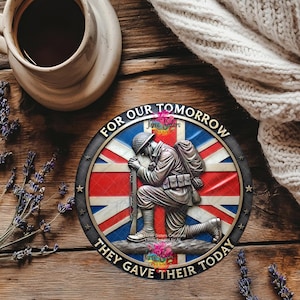 May include: A round metal emblem featuring a kneeling soldier in front of a Union Jack flag. The text "FOR OUR TOMORROW THEY GAVE THEIR TODAY" encircles the design. The emblem is set on a wooden surface with a cup of coffee and lavender.