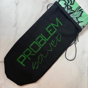 Puede incluir: Bolsa negra con cordón con las palabras "PROBLEM SOLVED" en letras verdes brillantes. La parte superior de la bolsa es de tela verde claro con un diseño floral negro. La bolsa está sobre una superficie de mármol blanco y gris.