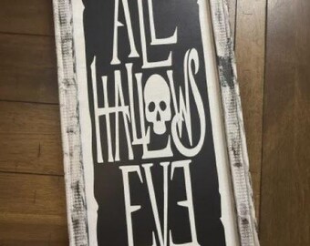 Op de afbeelding: Rechthoekig houten bord met de woorden "ALL HALLOWS EVE" in het wit, met een schedelafbeelding. De achtergrond is zwart en de lijst is wit met een verweerde afwerking. Een Halloween-decoratie.