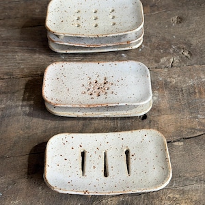 May include: A collection of rectangular ceramic soap dishes with a speckled off-white glaze and brown edges. Each dish features distinct drainage patterns, including circular holes and vertical slits, designed to hold soap bars securely.