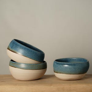 May include: Four ceramic bowls with a two-tone design. The top half of each bowl is a deep blue, while the bottom half is a creamy white. The bowls are stacked and arranged on a wooden surface.