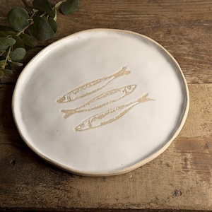 May include: A round, white ceramic plate with an irregular, hand-formed edge. The centre of the plate features a debossed, light brown graphic design of three fish arranged diagonally, one above the other, with a smooth, glossy finish.