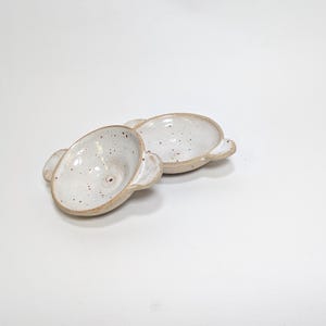 May include: Two handmade ceramic dishes. Each oval dish is white with brown speckles and small handles. The dishes are unglazed on the bottom, with a rustic, organic aesthetic. The dishes are likely made in the UK.