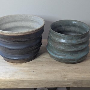 May include: Two handmade ceramic bowls with a ribbed design. One bowl is dark blue with a light brown rim, and the other is teal. Both bowls are on a light brown wooden surface.
