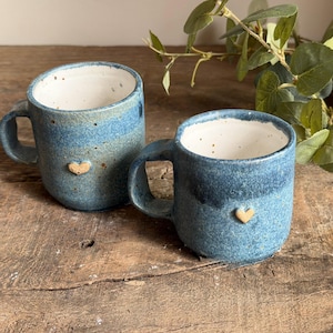 May include: Two handmade ceramic mugs with a speckled blue glaze and a white interior. Each mug features a small, raised, light-coloured heart emblem on the front exterior. The mugs have a rounded shape with a sturdy handle on the side.