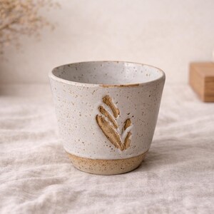 May include: A speckled ceramic mug with a leaf design. The off-white mug has a textured surface and a light brown base. The leaf design is carved into the mug's surface. The mug is a handmade item.