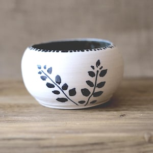 May include: A white ceramic bowl with a black interior and a decorative black leaf design. The bowl is round and sits on a wooden surface. The rim of the bowl has a dotted pattern.