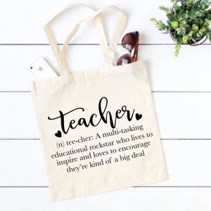 Peut inclure: Un sac fourre-tout blanc avec du texte noir qui dit "teacher" et une d&eacute;finition du mot. La d&eacute;finition se lit comme suit : "(n) tee-cher : Une rockstar multit&acirc;che de l'&eacute;ducation qui vit pour inspirer et aime encourager, c'est un peu un gros probl&egrave;me."