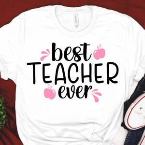 Peut inclure: T-shirt blanc avec le texte "best teacher ever" en lettres noires et un graphique de pomme rose.