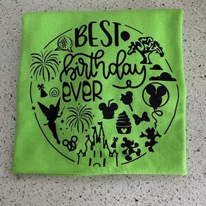 May include: A green t-shirt with a black design that says "BEST birthday EVER" surrounded by various Disney-themed icons, including Mickey Mouse, Tinkerbell, fireworks, and a castle.