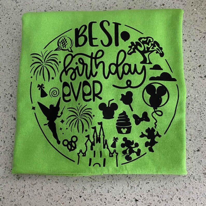 May include: A green t-shirt with a black design that says "BEST birthday EVER" surrounded by various Disney-themed icons, including Mickey Mouse, Tinkerbell, fireworks, and a castle.