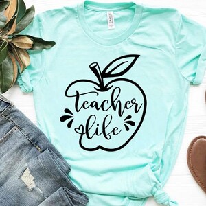 Peut inclure: Un t-shirt bleu clair avec un motif de pomme noir et le texte "teacher life" dans une police d&eacute;corative.