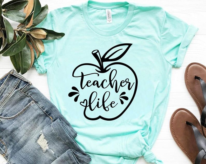 Peut inclure: Un t-shirt bleu clair avec un motif de pomme noir et le texte "teacher life" dans une police d&eacute;corative.