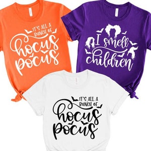 May include: Three t-shirts with Halloween-themed designs. One orange shirt says "It's all a bunch of Hocus Pocus" with bats and a witch's hat. One purple shirt says "I smell children" with bats and a witch's hair. One white shirt says "It's all a bunch of Hocus Pocus" with bats.