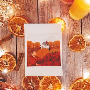 Peut inclure: Une carte blanche avec une illustration peinte à la main d'une personne cueillant des citrouilles devant une petite maison. La carte est entourée de tranches d'orange séchées, de bâtonnets de cannelle et de guirlandes lumineuses blanches sur une surface en bois.