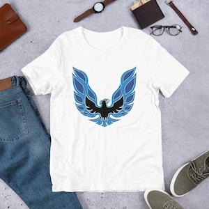 Puede incluir: Camiseta blanca con un logotipo de fénix azul y negro. El logotipo representa un pájaro estilizado con las alas extendidas.
