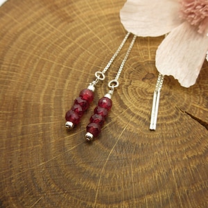 Pode incluir: Um par de brincos de prata com uma corrente pendente e contas de pedras preciosas vermelhas. Os brincos são exibidos em uma superfície de madeira com uma flor rosa.