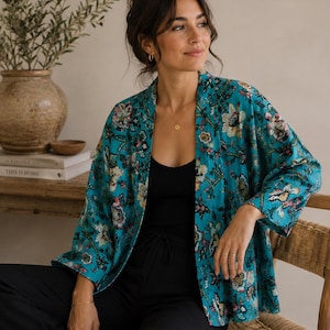 May include: A teal open-front jacket featuring a vibrant floral pattern in shades of pink, white, and green. The garment has long, loose sleeves and a relaxed fit, worn over a simple black vest top and black trousers to complete the ensemble.
