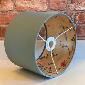 May include: A sage green lampshade with a floral and bird print lining. The lampshade has a white fabric lining and a white metal ring for attaching to a lamp base.
