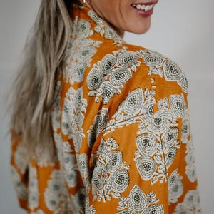 May include: Mustard-colored button-down shirt with a white and dark green paisley pattern. The shirt is made of a lightweight fabric and has long sleeves. The image shows the back of the shirt.