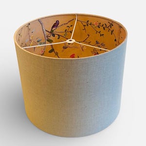 May include: A cylindrical lampshade with a light blue exterior and a yellow interior. The interior features a detailed floral and bird design. The lampshade has a white metal frame and is suitable for a table or floor lamp.