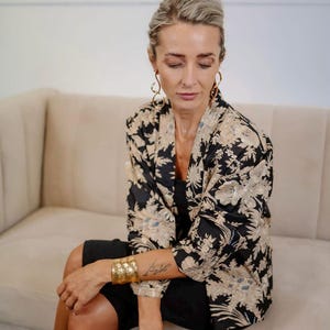 Op de afbeelding: Een zwarte jas in kimono-stijl met een bloemenpatroon in beige en wit. De jas wordt over een zwarte jurk gedragen. Een gouden manchetarmband en grote oorringen maken de look compleet. De persoon zit op een beige bank.