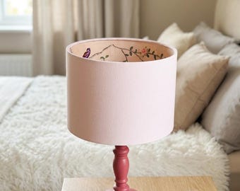 Op de afbeelding: Een tafellamp met een effen roze cilindrische kap en een donkerroze gedraaide houten voet. De binnenkant van de lampenkap is voorzien van een printpatroon van dunne bruine takken, kleine groene blaadjes, roze bloemen en een kleine paarse vogel.