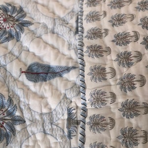 May include: A white cotton quilt with a blue and white floral pattern. The quilt is quilted in a diamond pattern and has a white border. The quilt is made of two different fabrics, one with a floral pattern and the other with a tree pattern.