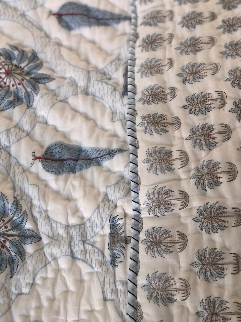 May include: A white cotton quilt with a blue and white floral pattern. The quilt is quilted in a diamond pattern and has a white border. The quilt is made of two different fabrics, one with a floral pattern and the other with a tree pattern.