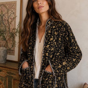 May include: A black quilted jacket featuring a gold floral pattern, a round neckline, and two front pockets. The jacket is finished with a black and white striped trim along the front opening, the neckline, the sleeve cuffs, and the pocket edges.