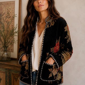 May include: A black velvet jacket featuring a floral pattern in shades of red, green, and gold. The jacket has a stand-up collar, long sleeves, and two front patch pockets. The edges of the collar, front opening, and pockets are trimmed with white stitching.