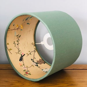 May include: A green lampshade with a floral and bird design on the inside. The lampshade is made of fabric and has a white ring at the top.