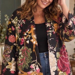 May include: A black open-front jacket featuring a vibrant botanical print with large red, pink, yellow, and green floral and leaf patterns. The garment has long, wide sleeves and a straight hemline, worn over a black top and blue denim jeans.