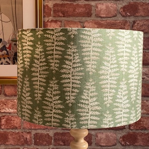 May include: A green lampshade with a white fern design. The lampshade is cylindrical and rests on a light-coloured wooden lamp base. The background includes a red brick wall and a framed picture.