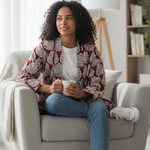 Puede incluir: Un kimono con estampado burdeos y crema. La prenda se lleva sobre una camiseta blanca y unos vaqueros azules. La persona está sentada en un sillón gris, sosteniendo una taza blanca. Ambiente relajado.