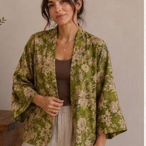 Op de afbeelding: Een olijfgroen kimono-jasje met open voorzijde en een herhalend bloemenpatroon in een lichtbeige tint. Het kledingstuk heeft wijde driekwartmouwen en een rechte, relaxte pasvorm die losjes over een effen bruine tanktop wordt gedragen.