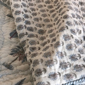 May include: A white cotton quilt with a blue and brown palm tree print. The quilt is folded in half and has a blue trim around the edges.