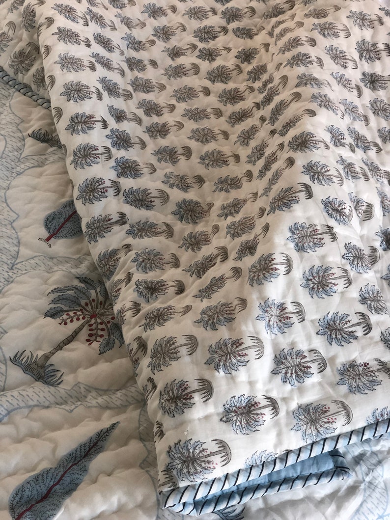 May include: A white cotton quilt with a blue and brown palm tree print. The quilt is folded in half and has a blue trim around the edges.