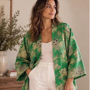 May include: A green open-front jacket featuring a floral pattern in shades of gold and cream. The garment has wide, three-quarter length sleeves and a straight hemline, worn over a plain white top and white trousers.