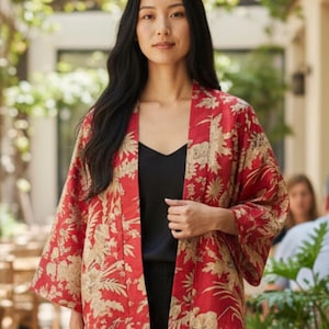 Puede incluir: Chaqueta tipo kimono roja con un estampado floral dorado. La chaqueta abierta tiene mangas anchas. La modelo lleva un top negro y pantalones negros. El fondo es un entorno exterior borroso con vegetación.