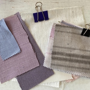 May include: A selection of fabric swatches in shades of lavender, pale blue, and cream. The samples are secured with binder clips. Some feature striped designs, and all have a textured finish, possibly linen or a similar fabric. Measurements are not available.