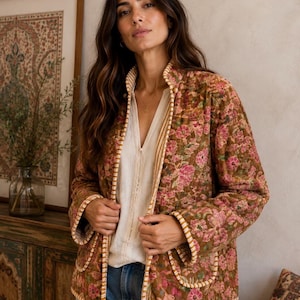 May include: A quilted jacket featuring a brown base with a pink and green floral pattern, a high collar, and white and brown striped trim along the edges and pockets. The jacket is worn over a cream-coloured button-down shirt and blue denim jeans.