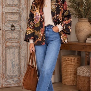 May include: A black open-front jacket featuring a multi-colored floral and botanical print, worn over a cream-colored knit sweater and blue denim jeans. The outfit is paired with brown suede ankle boots and a matching brown suede tote bag.