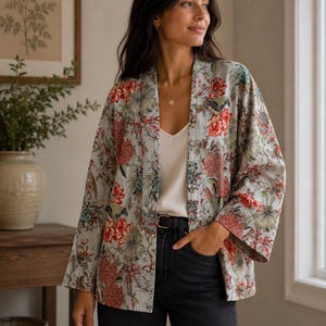 May include: A light grey open-front jacket featuring a vibrant floral print with red, green, and blue botanical patterns. The garment has wide, three-quarter length sleeves and a straight hemline, worn over a simple white camisole and black jeans.