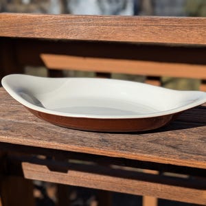 May include: An oval serving dish with a white interior and a brown exterior. The dish has a smooth, glossy finish and is on a wooden surface. The dish is empty and appears to be ceramic.