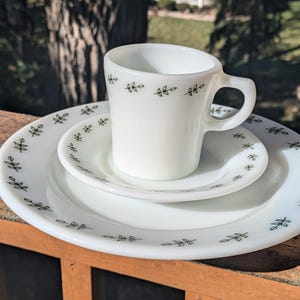 May include: A white ceramic cup and saucer set with a green leaf pattern. The cup has a handle and the saucer is slightly larger than the cup.