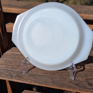 May include: A white, oval-shaped Pyrex baking dish with a rounded center and angular sides. The dish has two handles and is displayed on a clear plastic stand. The dish is made of opaque white glass.