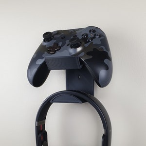 May include: A black and gray camouflage Xbox controller and black headphones are mounted on a black wall mount.