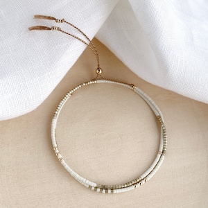 May include: A white and gold beaded bracelet with a sliding knot closure. The bracelet is made of small, round beads and has a delicate, minimalist design.